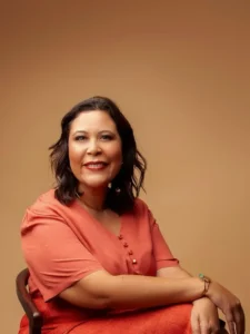 Mulher sentada sorrindo. Ela tem cabelo castanho escuro, na altura dos ombros, levemente ondulado. Está usando uma blusa de manga curta na cor salmão e uma saia ou calça em tonalidade semelhante. Usa brincos pequenos, colar fino e pulseiras, incluindo uma pulseira dourada com detalhe em verde. O fundo é liso e bege, criando uma sensação de harmonia com as cores da roupa. Ela está sentada de lado, mas voltada para a câmera, transmitindo simpatia e tranquilidade.