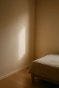 Quarto minimalista. A imagem mostra um canto de um quarto com estilo minimalista. No lado direito inferior, há uma cama simples com lençol bege claro cobrindo o colchão. As paredes são lisas, de cor bege clara, sem qualquer decoração. Um feixe de luz solar entra pela janela (fora do enquadramento), projetando um retângulo de luz suave na parede, criando uma atmosfera tranquila e aconchegante. O piso é de madeira clara, e não há outros móveis ou objetos visíveis no ambiente.