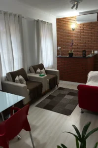 Sala de estar aconchegante com parede de tijolos aparentes ao fundo. No canto direito superior há um aparelho de ar-condicionado e uma luminária de parede acesa. Duas poltronas bege com cobertor marrom e almofadas claras estão posicionadas lado a lado, com uma mesa de apoio pequena entre elas segurando uma caixa de lenços. Em frente, há um tapete em tons de marrom com padrão quadriculado. À direita aparece parte de um sofá vermelho. À esquerda, mesa de vidro com cadeira vermelha. Sobre o balcão de tijolos há uma orquídea em vaso preto, uma luminária de mesa e alguns objetos pequenos. As janelas possuem cortinas brancas translúcidas, permitindo entrada de luz natural. O chão é claro, criando contraste harmonioso com os móveis.