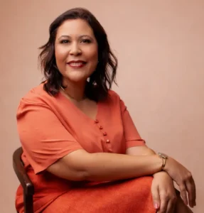 Mulher sentada em uma cadeira de madeira com estofado claro, posando para a foto diante de um fundo liso em tom bege rosado. Ela tem cabelos castanhos escuros, médios e levemente ondulados, usa vestido ou conjunto em tom alaranjado, sorrindo para a câmera. Os braços estão apoiados suavemente sobre as pernas cruzadas. Ela está usando acessórios discretos, como uma pulseira e brincos. O ambiente e a iluminação transmitem uma atmosfera acolhedora e profissional.